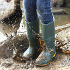 Splashing in a muddy puddle wearing Classic Wellies Green. The wellies have a bright yellow sole and the wearer has denim jeans on.